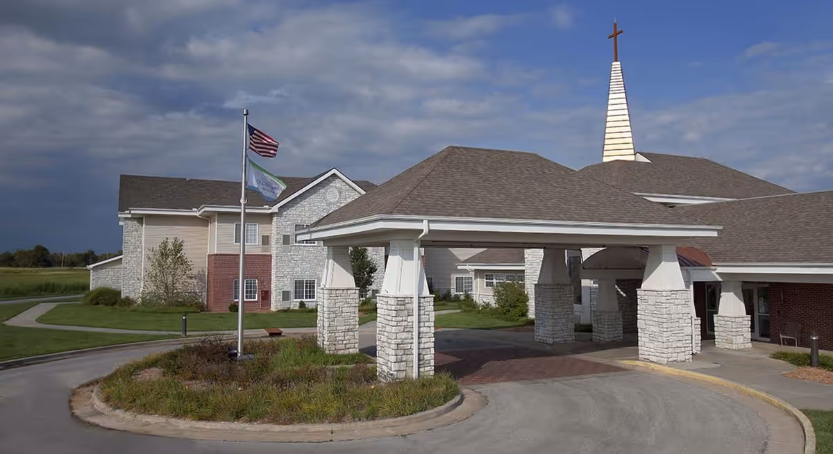 Exterior view of Ascension Living Via Christi Village showing the entrance with a covered drop-off area supported by stone pillars, an American flag and another flag on a flagpole, and a building with a tall steeple topped with a cross under a partly cloudy sky.