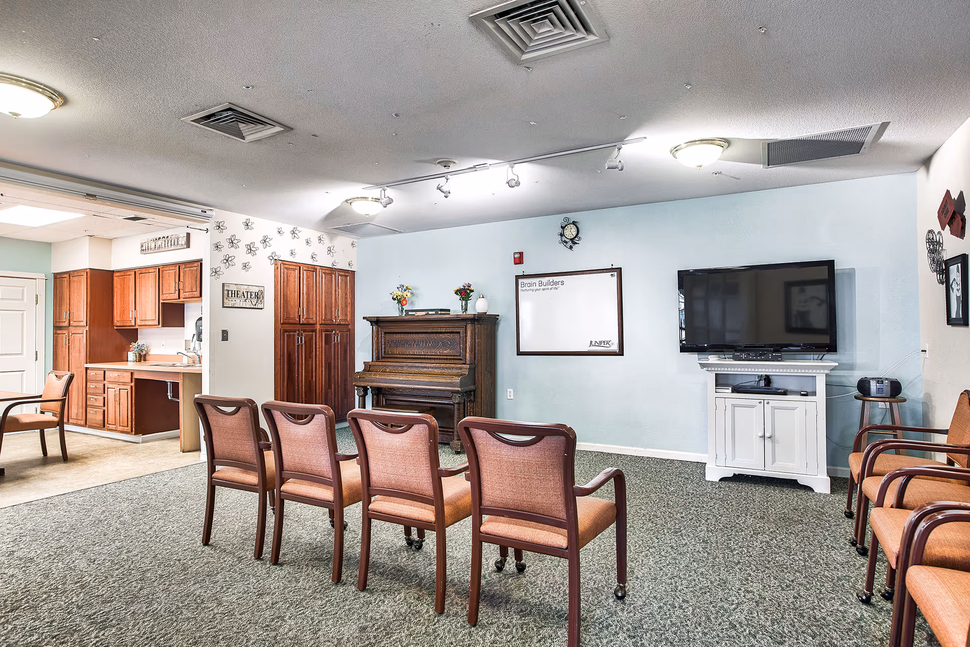 A community room with a row of chairs facing a large flat-screen TV mounted on a white cabinet. To the left, there is an upright piano with flowers on top and a whiteboard on the wall labeled 'Brain Builders'. The room has carpeted flooring and light blue walls, with an adjacent kitchen area visible through an open doorway.