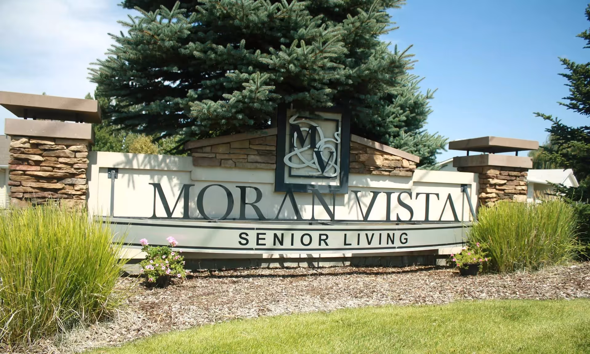 Stone entrance sign reading "MORAN VISTA SENIOR LIVING" surrounded by landscaping and evergreen trees.