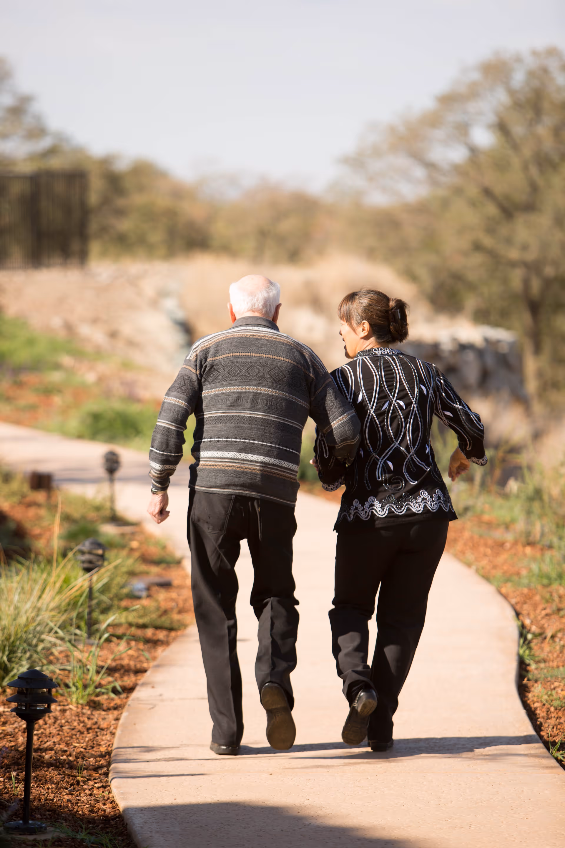 An elderly man and a woman walking arm in arm on a paved outdoor path surrounded by greenery and trees on a sunny day.