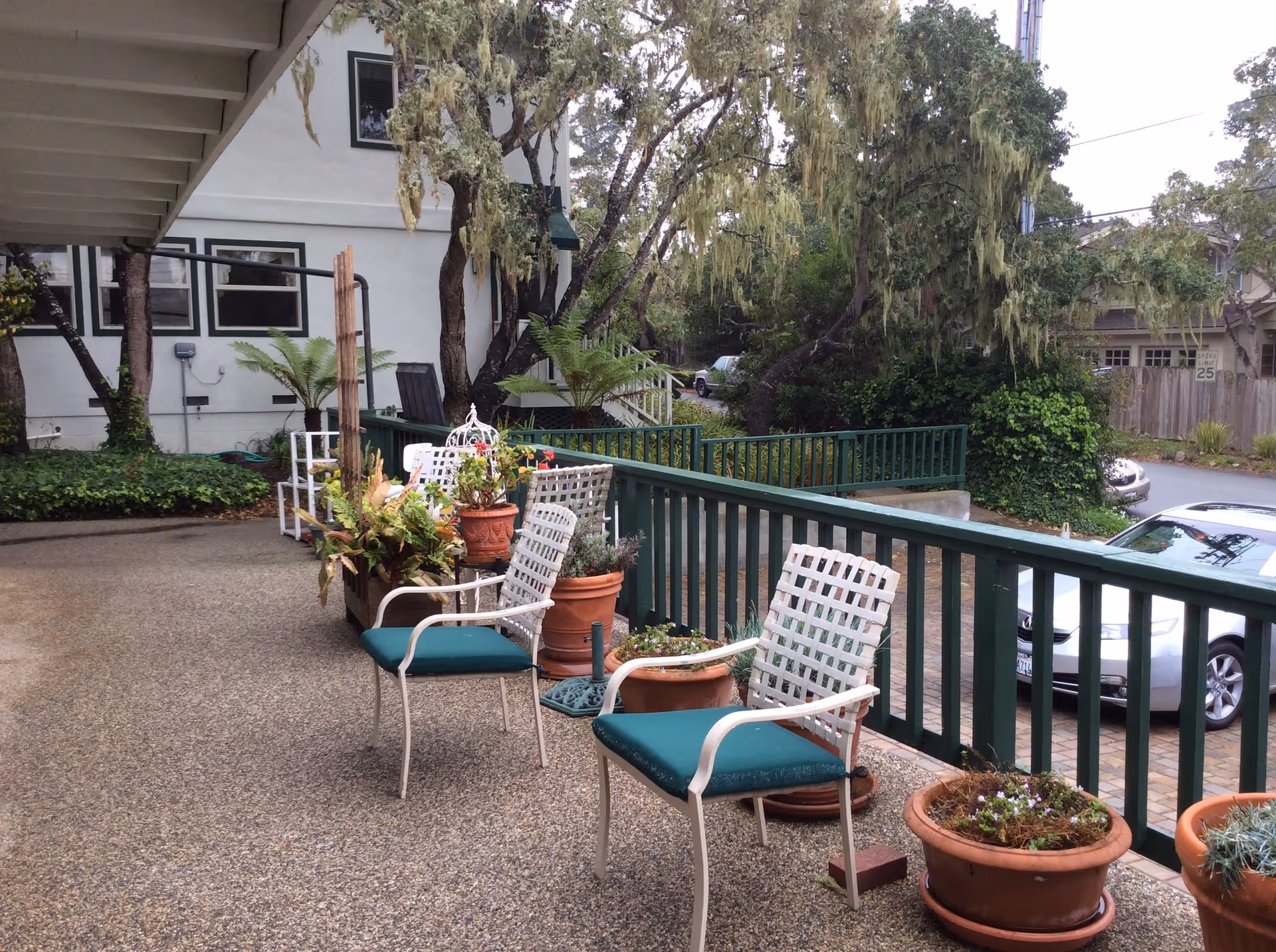 Outdoor patio area with green cushioned white metal chairs and several potted plants along a green railing. Trees and parked cars are visible in the background near a residential street.