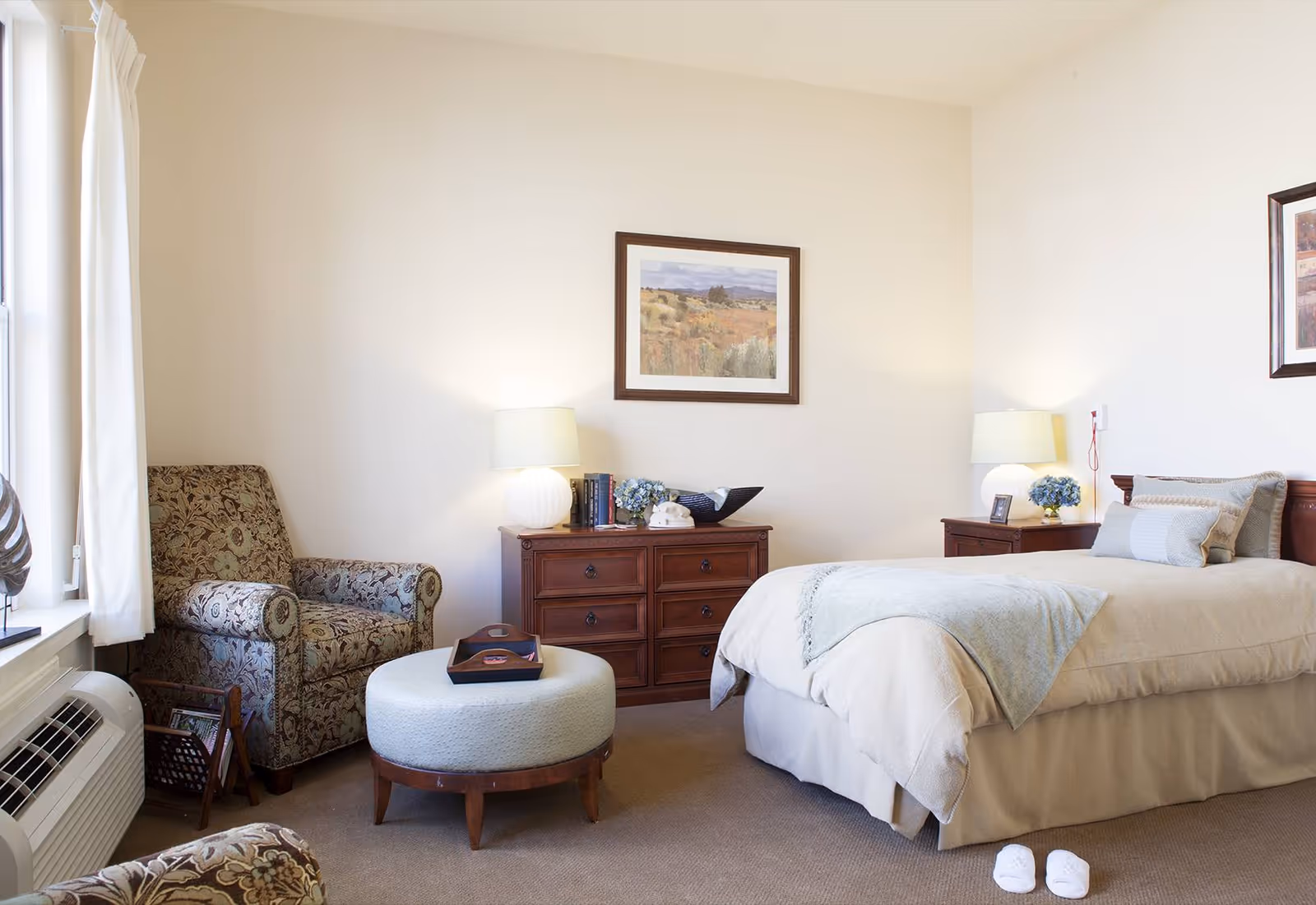 A cozy bedroom with a single bed covered in light-colored bedding and pillows, two wooden nightstands with lamps and decorative items, a wooden dresser with books and a decorative bowl, a patterned armchair near a window with white curtains, and a round upholstered ottoman with a tray on top.