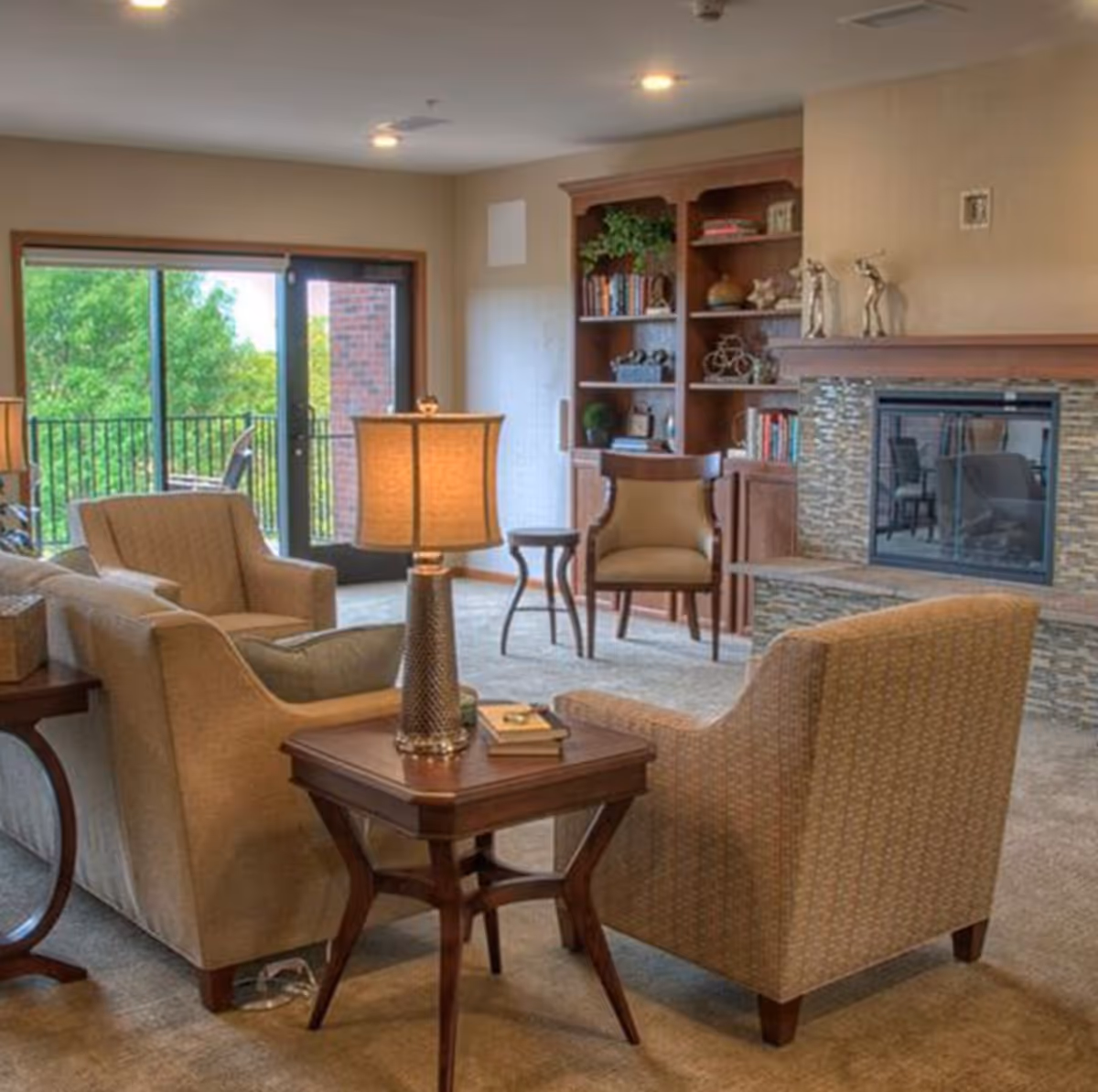 A cozy living room with beige upholstered armchairs and a sofa arranged around a wooden side table with a lamp and books. There is a stone fireplace with decorative sculptures on the mantel and a wooden built-in bookshelf filled with books and decor. Large sliding glass doors open to a balcony with a view of green trees outside.