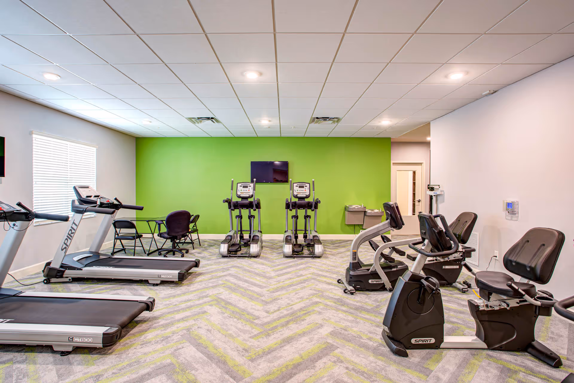 A bright fitness room with treadmills, ellipticals and recumbent bikes facing a green accent wall with a mounted TV.