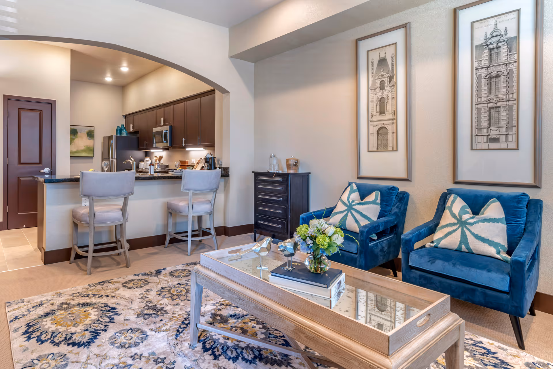 A cozy living area featuring two blue armchairs with patterned cushions, a wooden coffee table with a glass top holding decorative items and a flower arrangement, and a patterned area rug. In the background, there is an open kitchen with dark cabinets, a microwave, a refrigerator, and a breakfast bar with two cushioned stools. The walls are decorated with framed architectural prints.