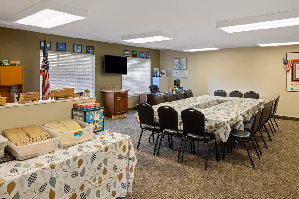 A meeting or activity room with a large rectangular table covered with a patterned tablecloth surrounded by black chairs. There is a smaller table in the foreground with trays of bingo tiles and boxes of party bingo. The room has beige walls, carpeted floor, two windows with blinds, an American flag, a wall-mounted TV, and framed pictures on the walls.