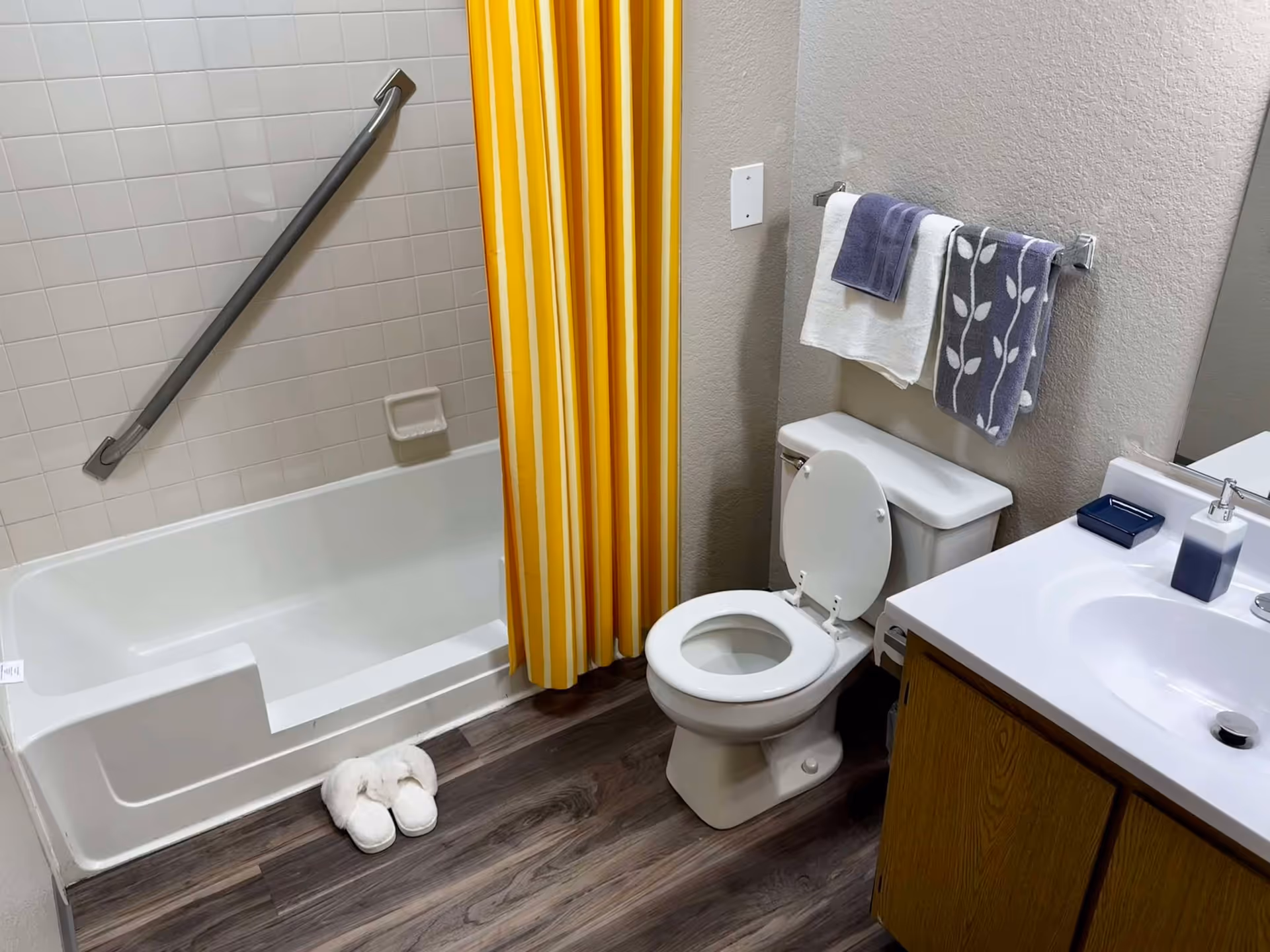 A clean bathroom featuring a white bathtub with a grab bar and yellow striped shower curtain, a white toilet with the seat up, a towel rack with three towels in white and purple colors, a wooden vanity with a white sink, soap dispenser, and a small blue container, and a pair of white slippers on the wooden floor.