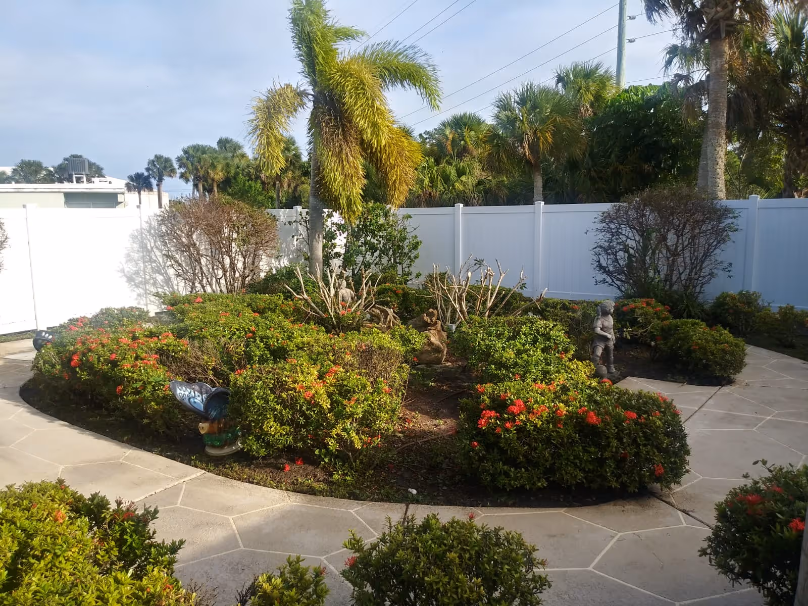 A landscaped outdoor garden area with a circular pathway surrounding green bushes with red flowers, palm trees, and small garden statues. A white fence encloses the garden, and the sky is clear with some clouds.