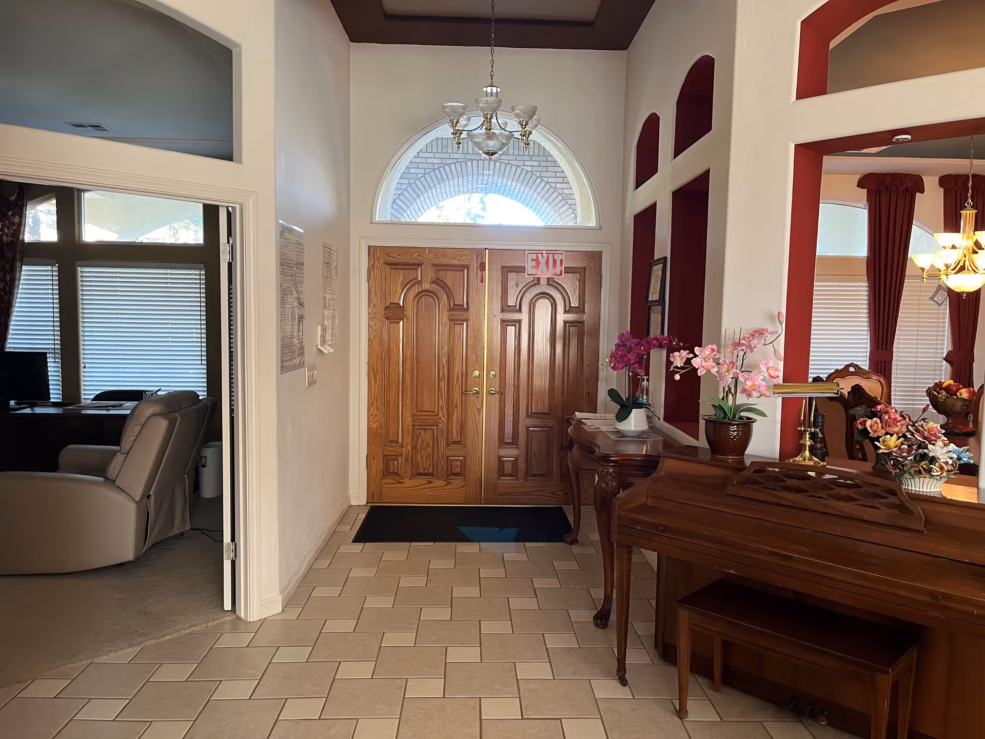 Entry foyer with double wooden doors, chandelier, a piano and floral arrangements opening into living and dining areas.