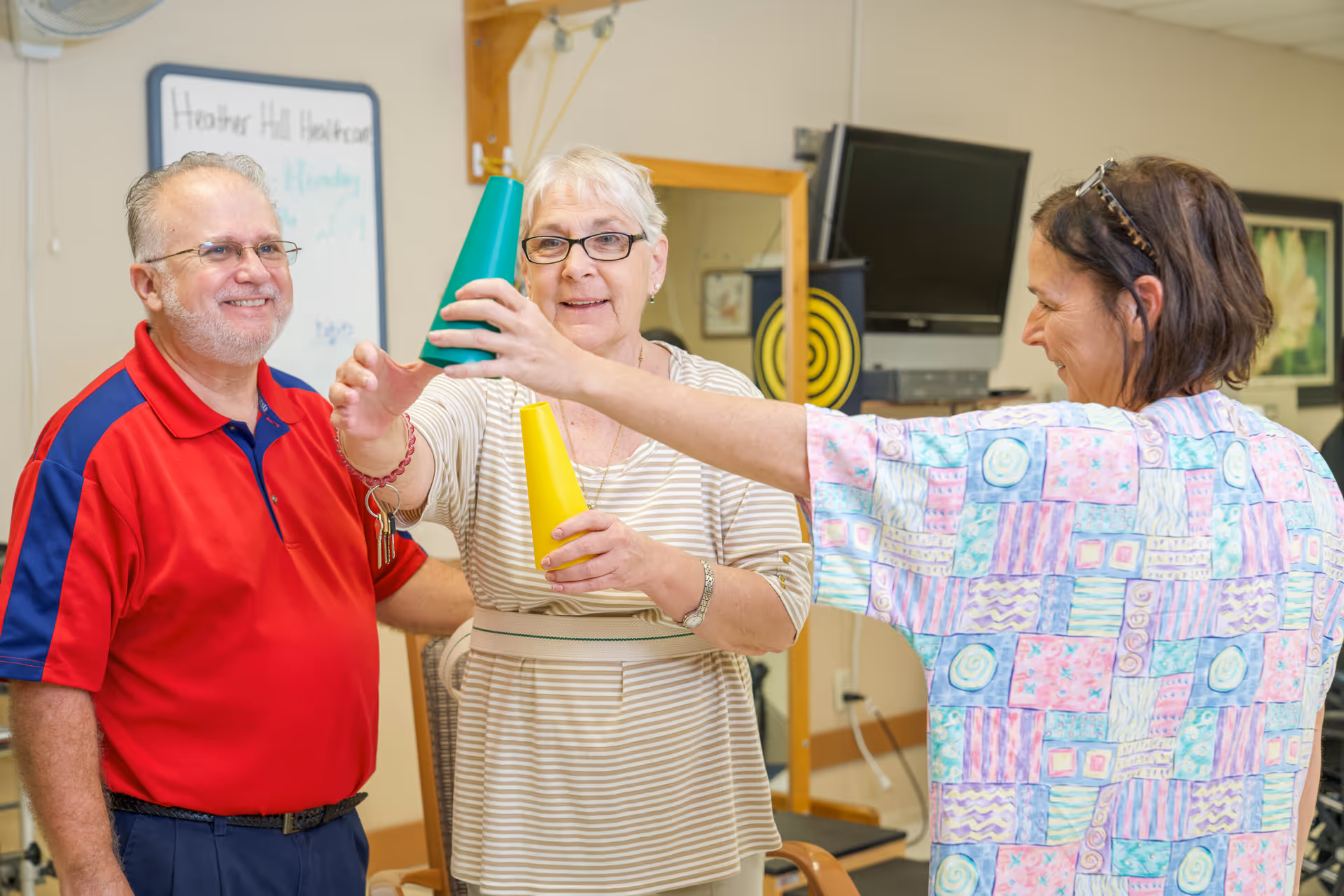 Three adults in a healthcare facility room engaging in a physical therapy or recreational activity involving stacking colorful cones. One woman is holding a yellow cone while another woman is placing a green cone on top. A man in a red shirt stands nearby smiling. A whiteboard with 'Heather Hill Healthcare' written on it is visible in the background.
