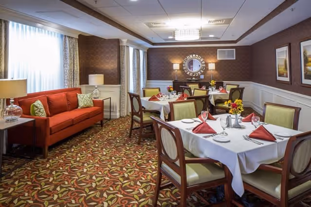 A dining room in a senior living facility with tables covered in white tablecloths, set with plates, glasses, silverware, and red folded napkins. The room has patterned carpet, a red sofa with green and white pillows, wall lamps, a round decorative mirror, and framed artwork on the walls.