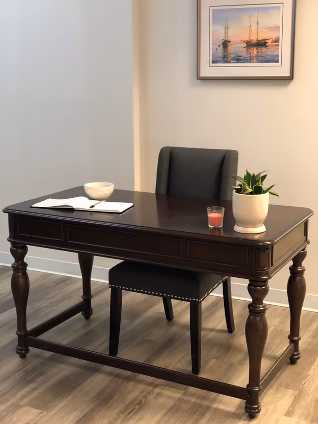 A dark wooden desk with turned legs is placed on a wood floor. On the desk are a white bowl, an open notebook with a pen, a small red candle, and a white pot with a green plant. Behind the desk is a dark upholstered chair with nailhead trim. A framed painting of sailboats on water at sunset hangs on the beige wall.