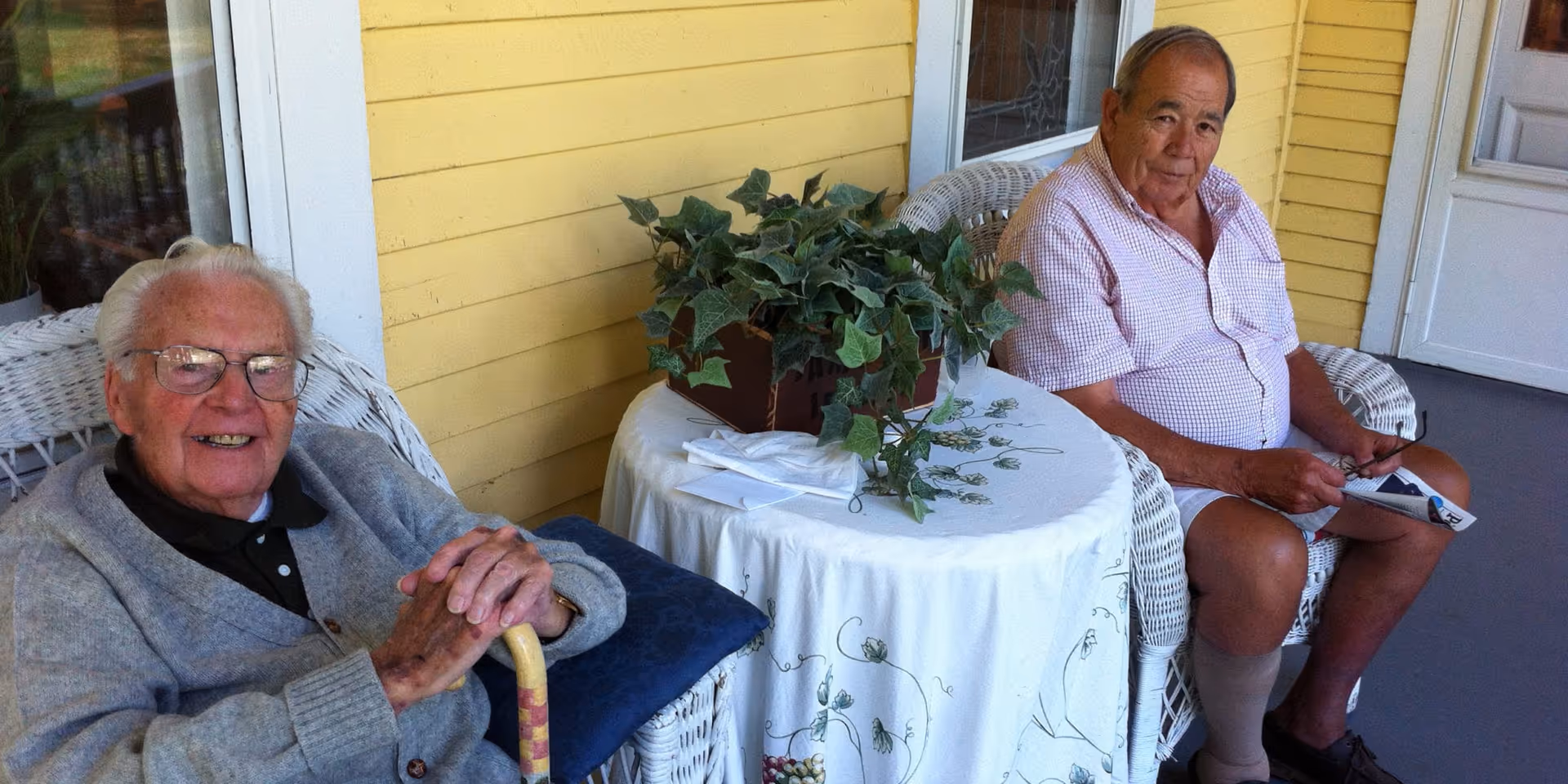 Two elderly men sit on a porch in wicker chairs beside a small round table with a potted plant.