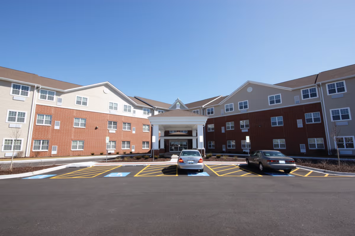 Exterior view of a senior living facility building with a covered entrance and two cars parked in designated handicap parking spaces in front. The building has a combination of brick and siding with multiple windows under a clear blue sky.