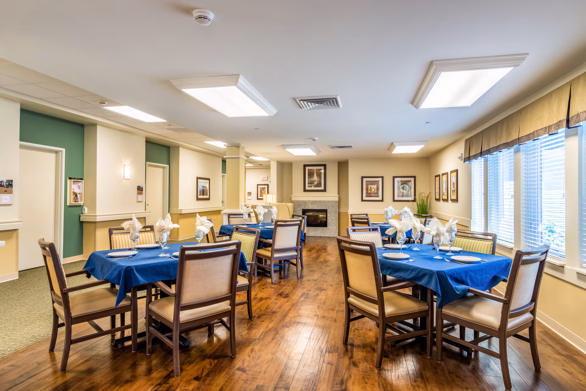 A dining room in Porter Place with several tables covered in blue tablecloths, each set with plates, glasses, and folded napkins. The room has wooden floors, beige walls with framed artwork, large windows with blinds and valances, and ceiling lights. There is a fireplace at the far end of the room.