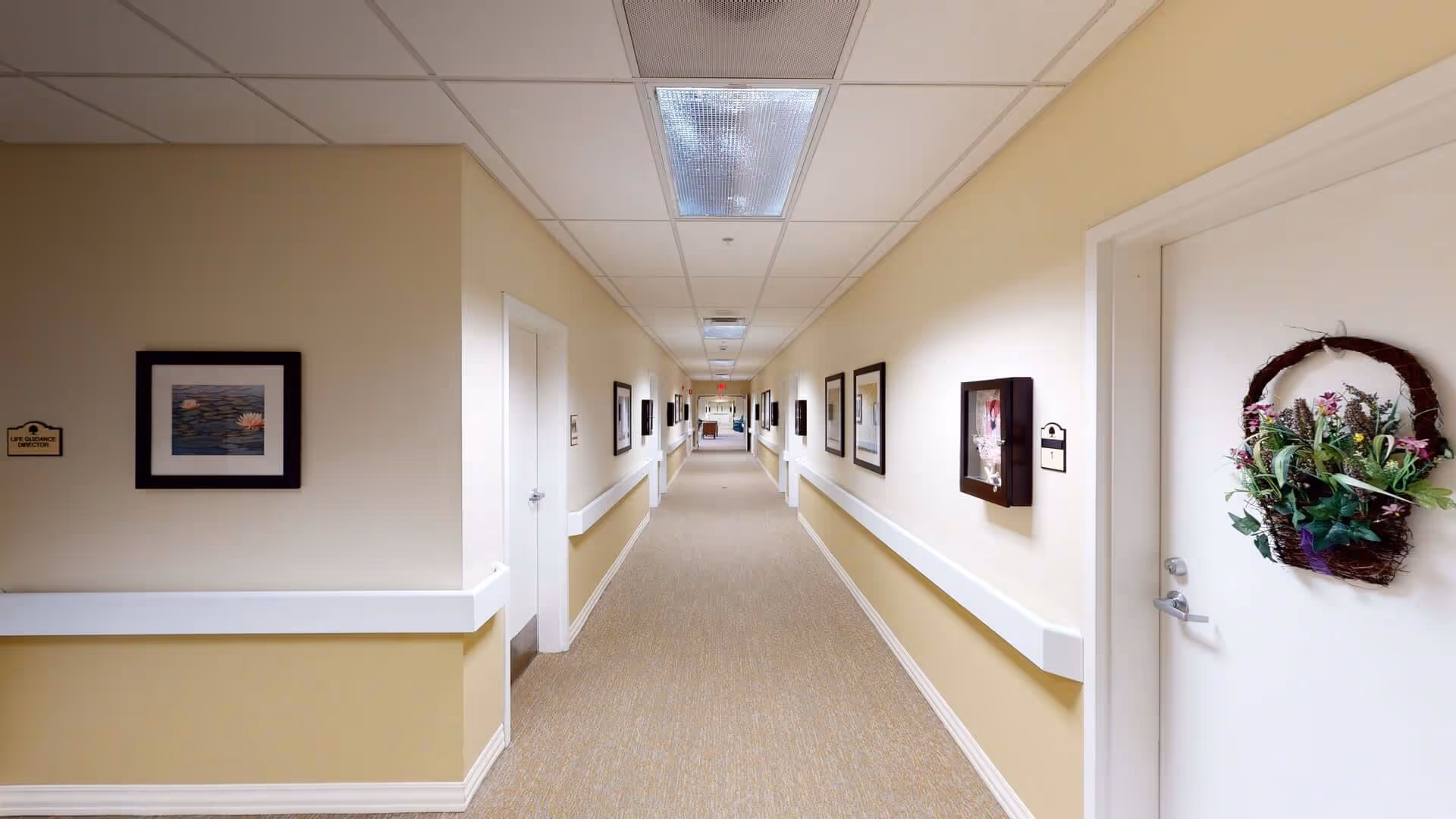 Long carpeted hallway inside a senior living facility with framed artwork and resident doors, one door decorated with a floral wreath.