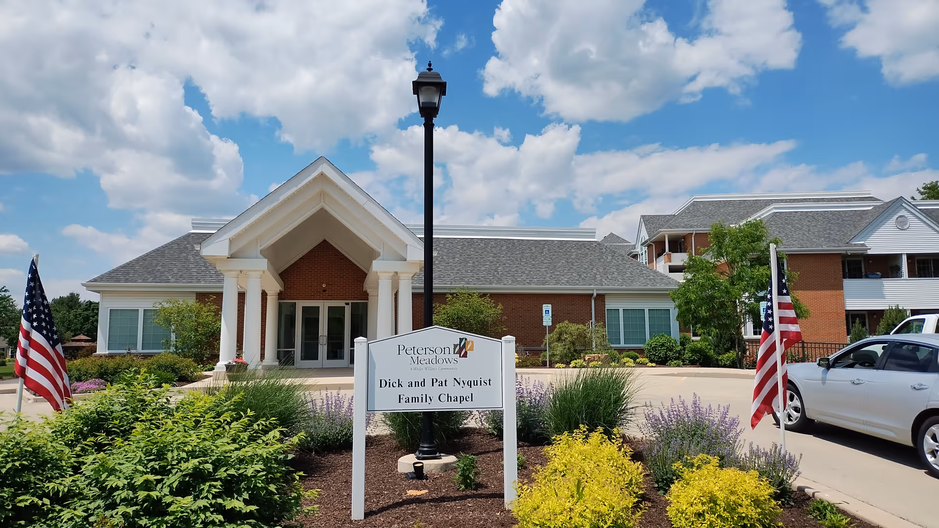 Exterior view of Peterson Meadows building with a sign in front that reads 'Dick and Pat Nyquist Family Chapel'. The building has a brick facade, white columns at the entrance, and a peaked roof. There are American flags on either side of the sign, bushes and flowers in the landscaped area, a streetlamp behind the sign, and a partly cloudy blue sky overhead.