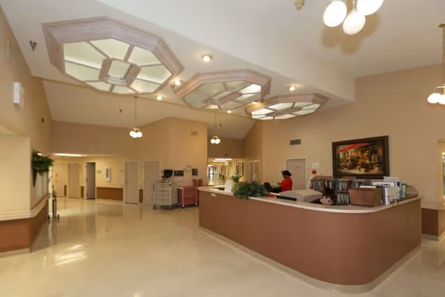 Spacious reception lobby with a curved front desk, decorative ceiling fixtures, and hallway entrances in a care facility.