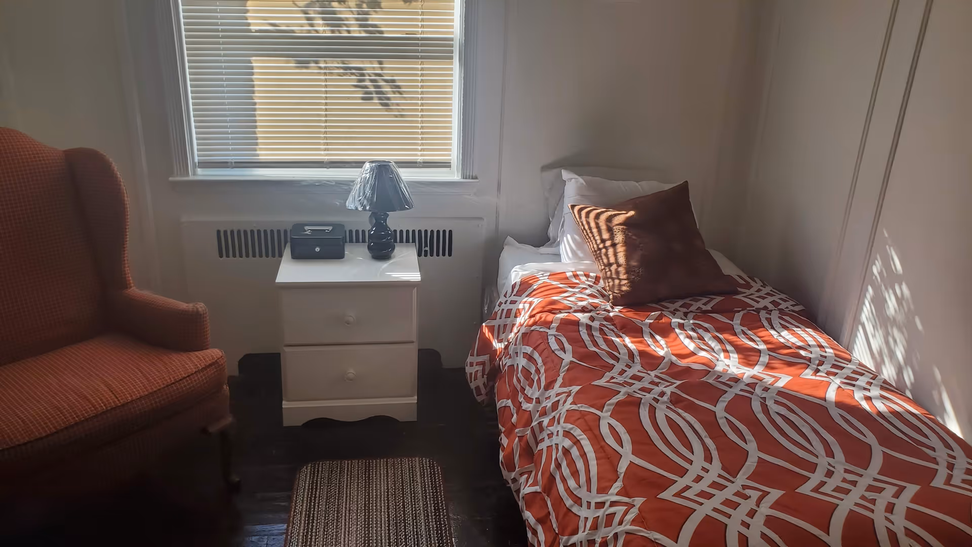 Sunlit small bedroom with a single bed covered in red-and-white patterned bedding, a white nightstand with a lamp under a window and an upholstered armchair.