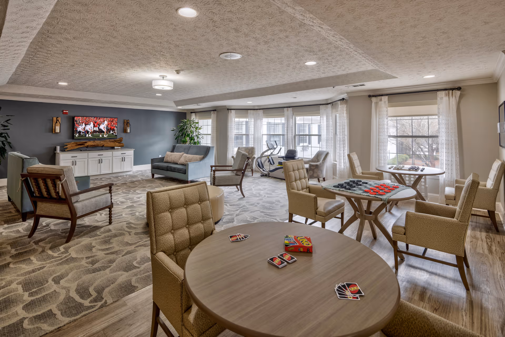 A spacious and well-lit common area in a senior living facility with multiple seating arrangements including armchairs and sofas. Several round tables are set up with board games and card games like checkers and Uno. Large windows with sheer curtains allow natural light to fill the room. A television is mounted on a dark accent wall above a white cabinet, and exercise equipment is visible near the windows.