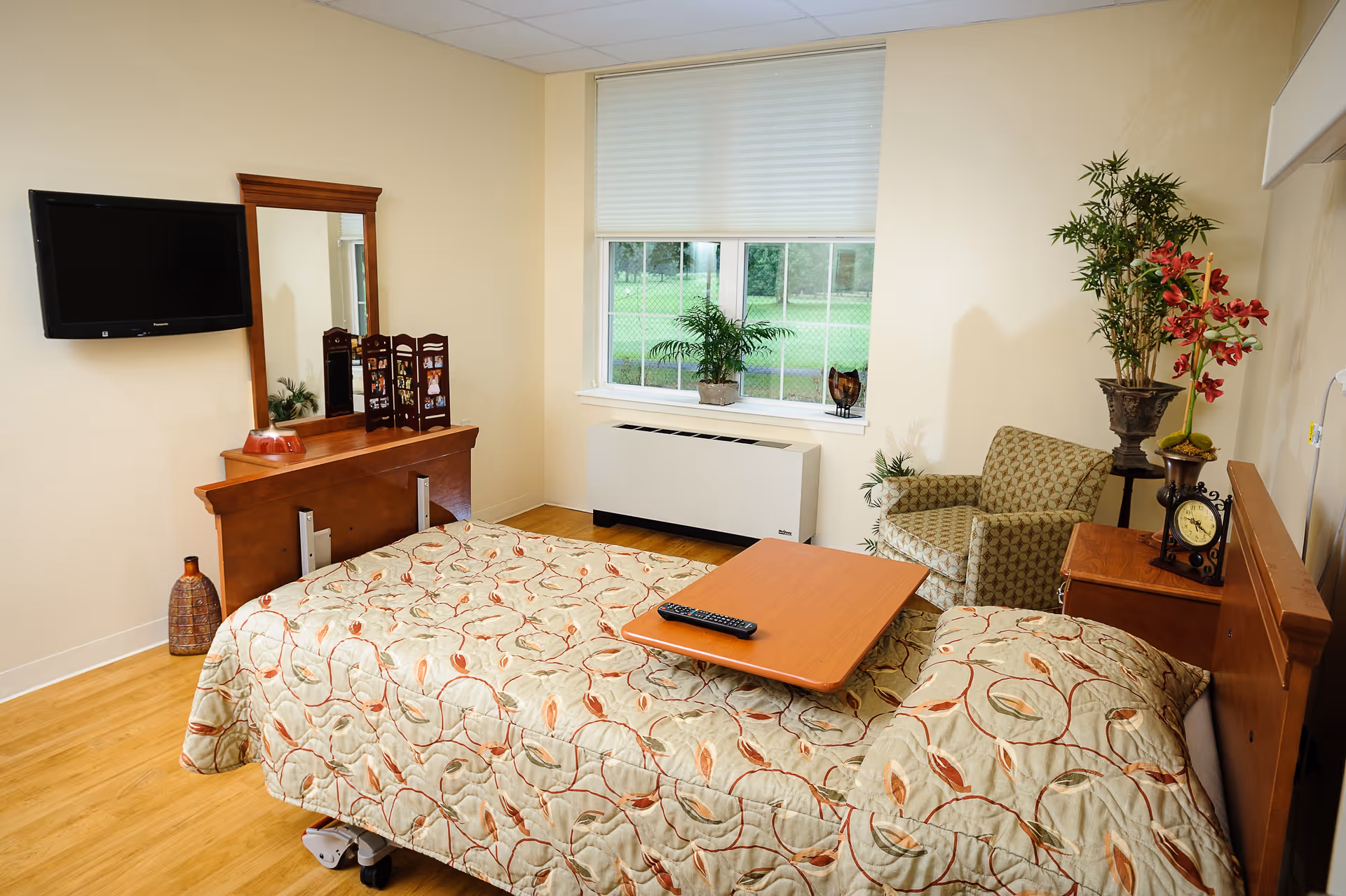 A nursing center bedroom with a single bed covered in a patterned quilt, a wooden headboard, and an overbed table with a remote control on it. The room has a wooden floor, a wall-mounted flat screen TV, a wooden dresser with a mirror, a small decorative folding screen, and a vase. There is a window with a view of green grass outside, a heating unit below the window, an armchair with a patterned fabric, and two potted plants on a side table and the floor.