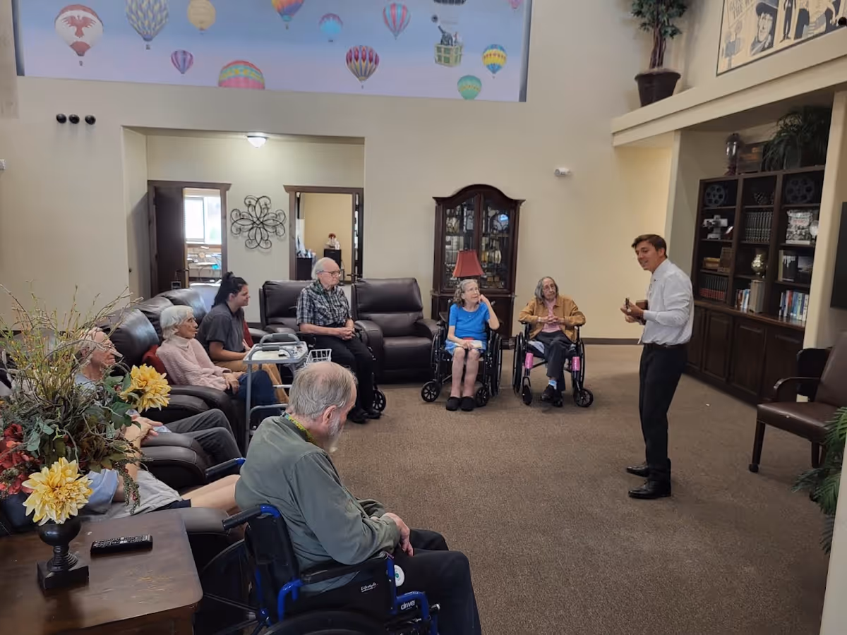 A group of elderly people, some in wheelchairs, seated in a semi-circle in a living room area of a senior living facility. A young man stands in front of them playing a small string instrument, possibly a ukulele. The room has a high ceiling with colorful hot air balloon decorations near the top, a wooden cabinet with glass doors, and bookshelves filled with books and decorative items. There are plants and a table with a flower arrangement in the foreground.