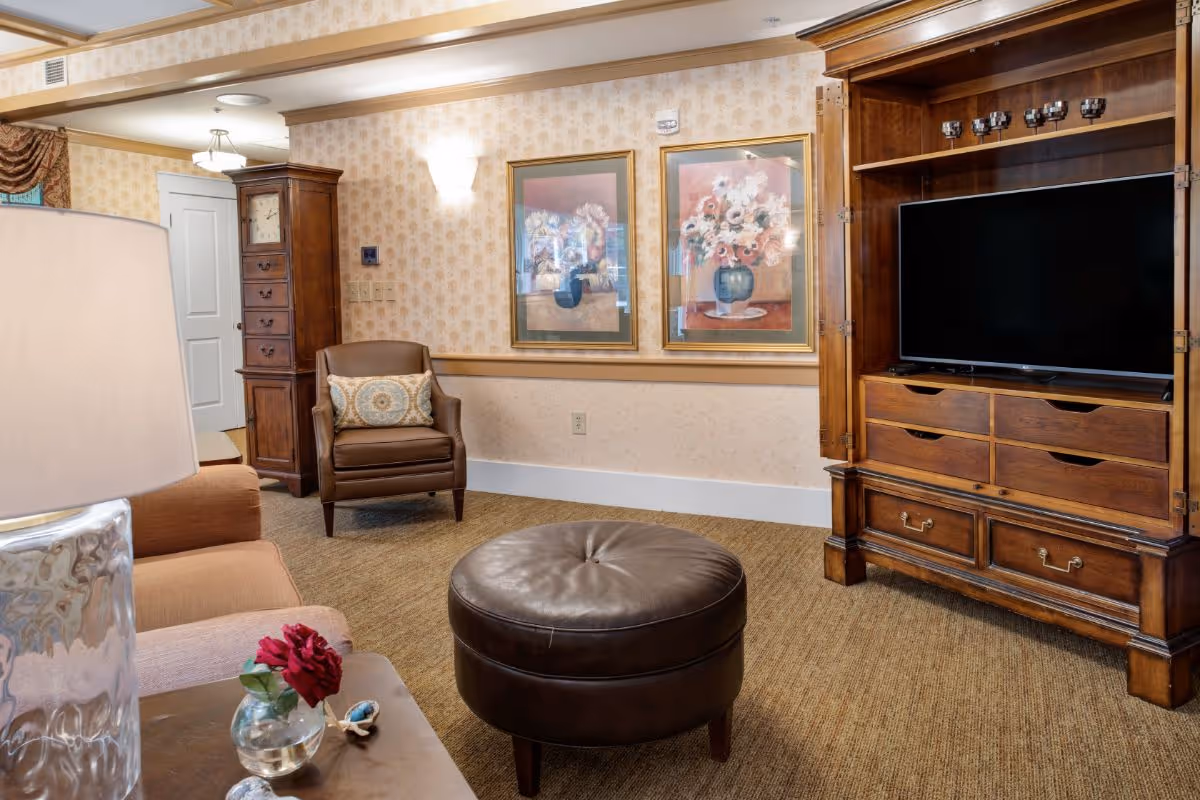A cozy living room area featuring a brown leather armchair with a decorative pillow, a round brown leather ottoman, and a wooden entertainment cabinet with a flat-screen TV. The walls are decorated with two framed floral paintings and patterned wallpaper. A table with a glass vase holding a red flower and a lamp with a textured glass base is partially visible in the foreground.