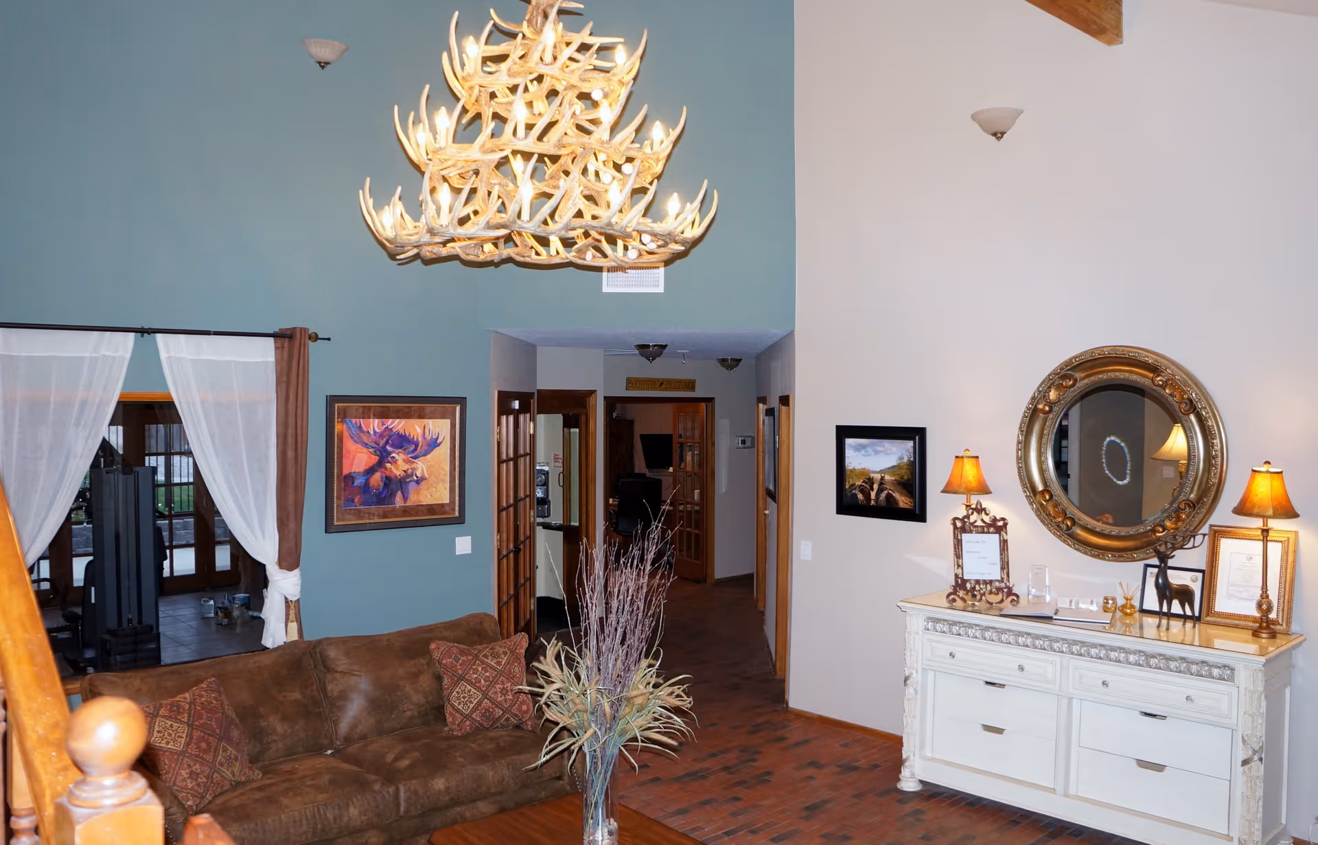 Spacious living room/entry with a brown leather sofa, antler chandelier, decorative console table with round mirror and lamps, and artwork on teal and beige walls.