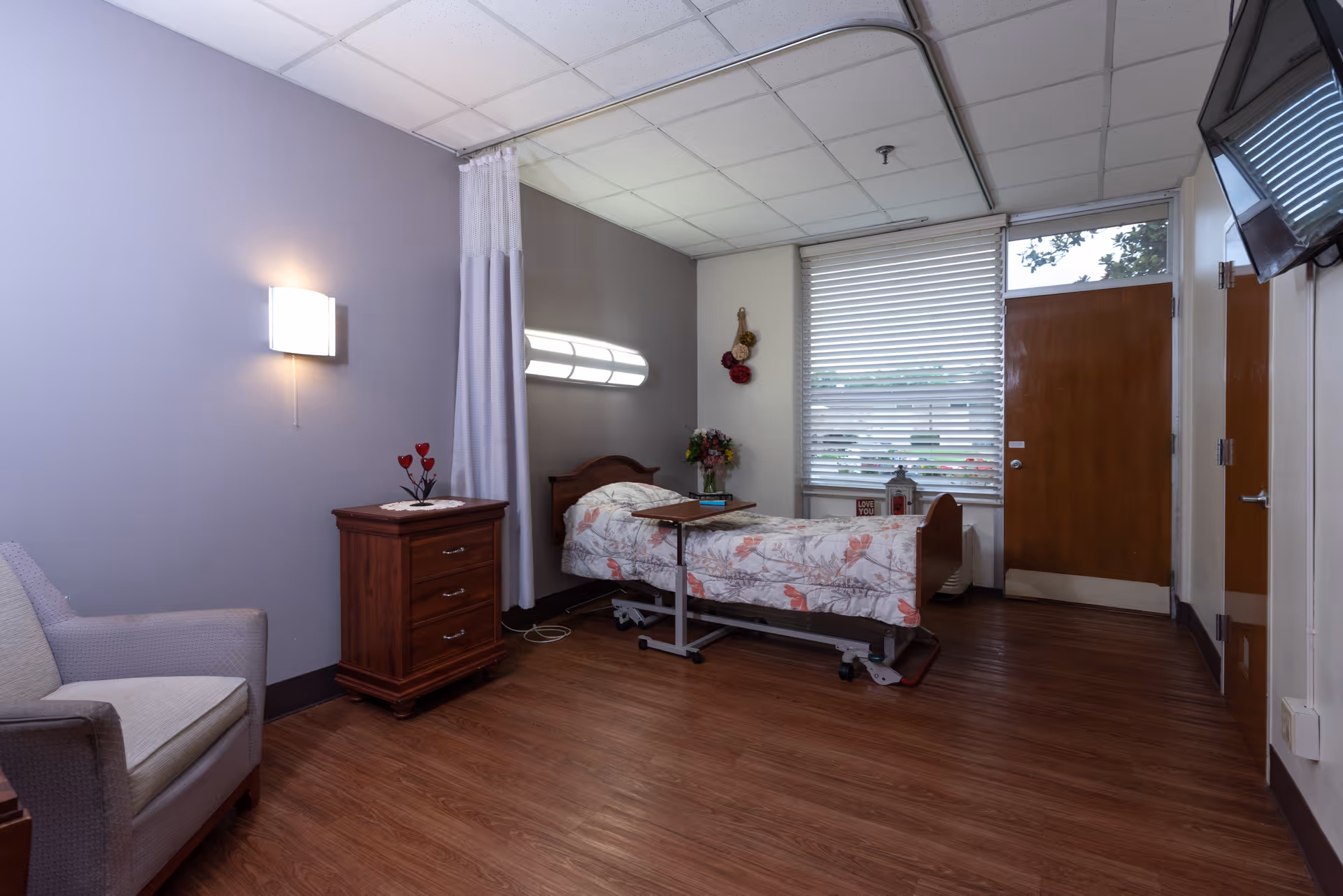 Private rehab patient room with a single adjustable bed, wooden nightstand, armchair, and a windowed door.