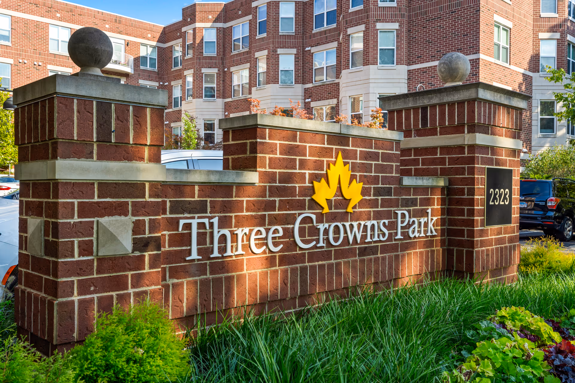 Brick entrance sign reading Three Crowns Park with a yellow crown logo and the apartment building behind it.