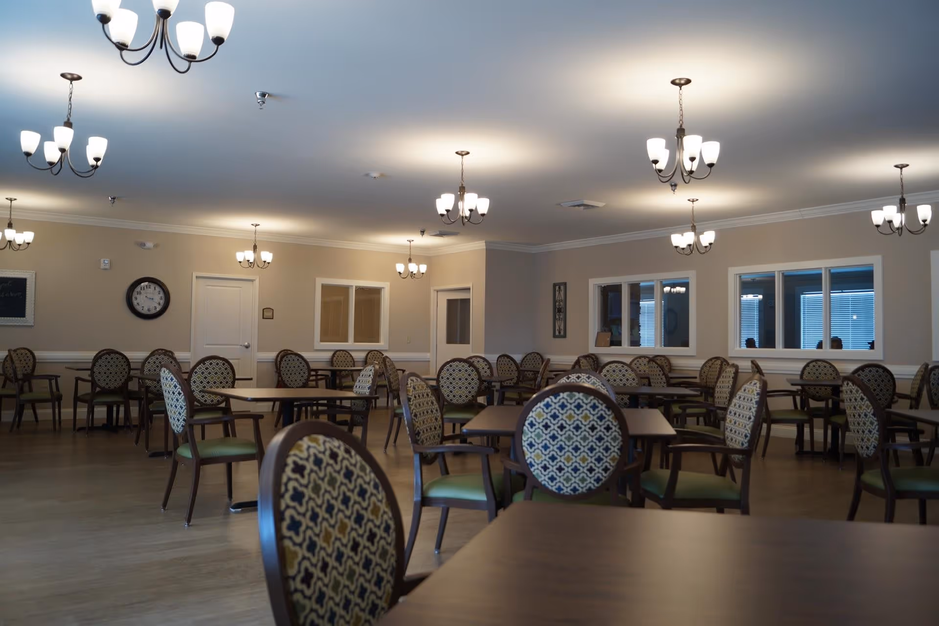 A spacious dining room with multiple tables and chairs featuring patterned upholstery. The room is well-lit with several chandeliers hanging from the ceiling. The walls are beige with white trim, and there are windows and doors visible along the walls.