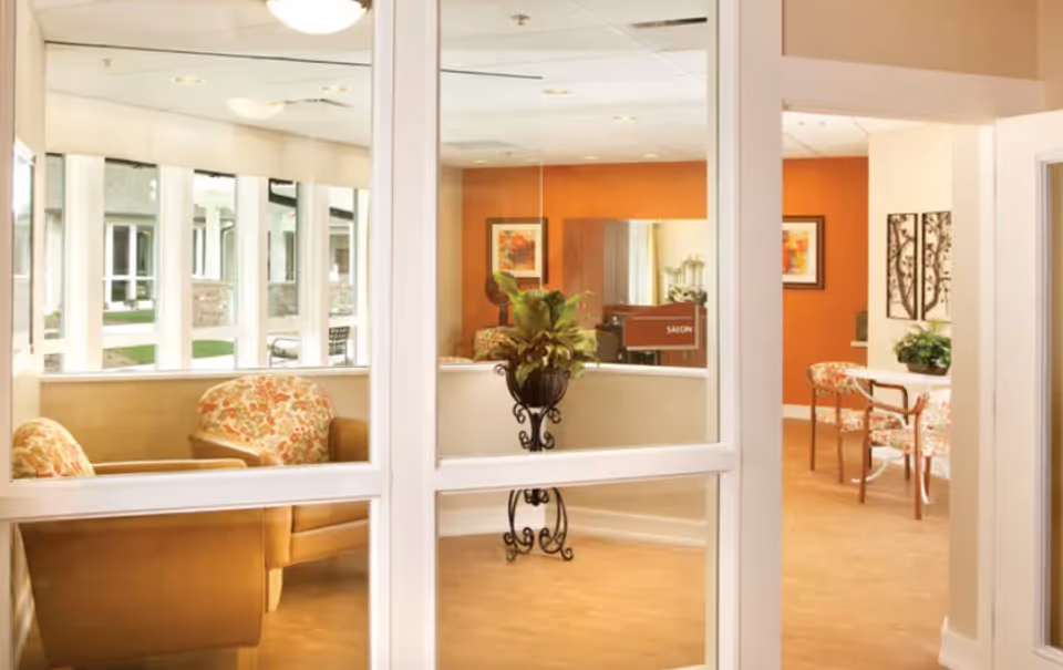 View through glass doors into a cozy seating area with floral upholstered chairs and a small table with chairs. The walls are painted orange and beige, decorated with framed artwork and a potted plant on a decorative stand. A sign indicating a salon is visible in the background.