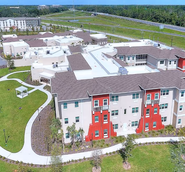 Aerial view of Promise Pointe Senior Living facility showing a large multi-wing building with beige and red exterior walls, surrounded by green lawns, landscaped gardens, and winding walking paths. The facility is located near a highway with trees and open land in the background.