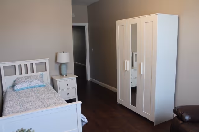 A simple, clean bedroom with a white single bed featuring patterned bedding and a pillow, a white nightstand with a blue lamp, a white wardrobe with a mirrored door, and a dark brown leather chair partially visible on the right. The room has beige walls and dark wooden flooring.