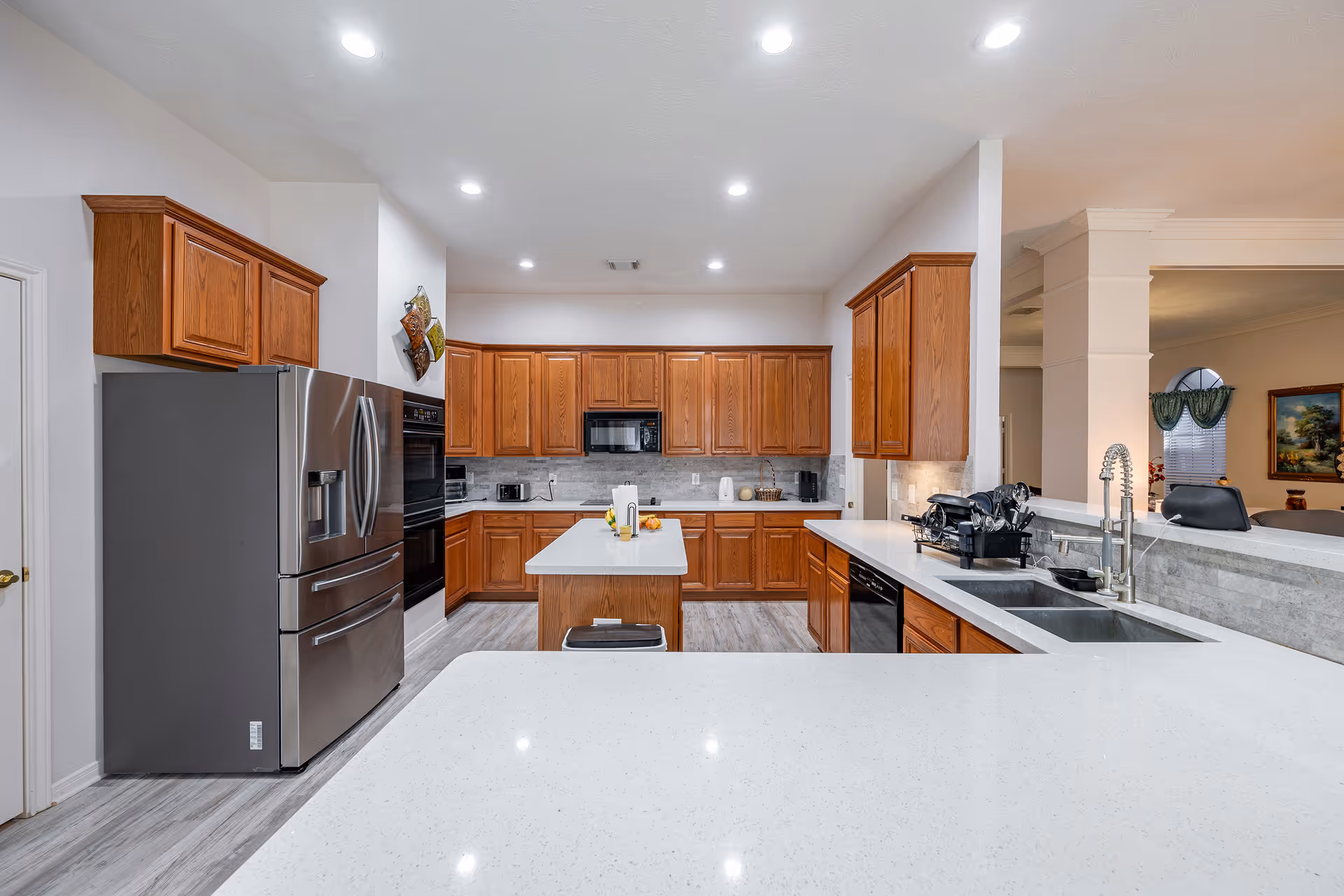 A spacious kitchen with wooden cabinets, a large stainless steel refrigerator, built-in oven, microwave, and a central island with a white countertop. The kitchen has a double sink, modern faucet, and various small appliances on the counters. The flooring is light-colored wood, and the kitchen opens into a living area with a window and framed artwork visible.