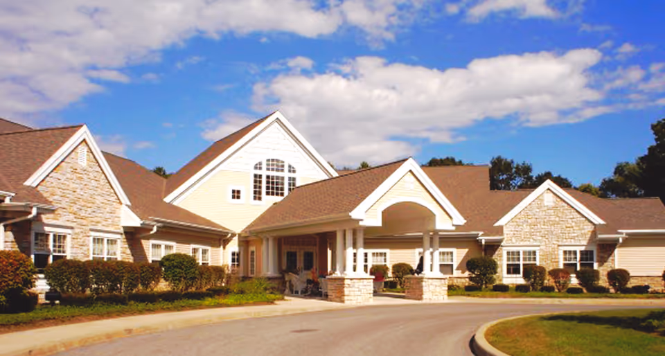 Exterior view of a single-story senior living facility building with beige siding and stone accents, a covered entrance supported by columns, surrounded by trimmed bushes and a curved driveway under a partly cloudy blue sky.