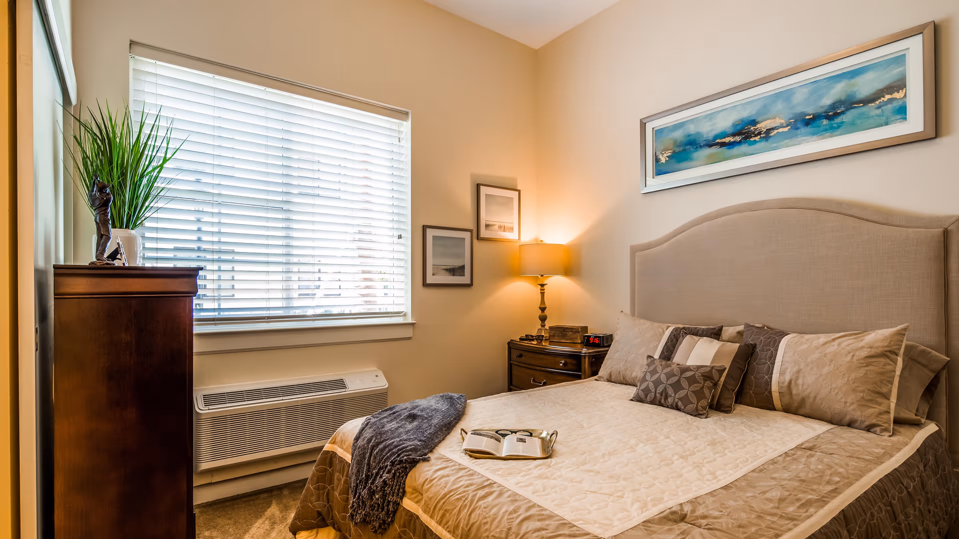 A cozy bedroom with a neatly made bed featuring beige and brown bedding and multiple pillows. There is a nightstand with a lamp and an alarm clock beside the bed. A tall wooden dresser with a potted plant and decorative statue is positioned near a window with white blinds. The walls are light-colored and decorated with framed artwork.