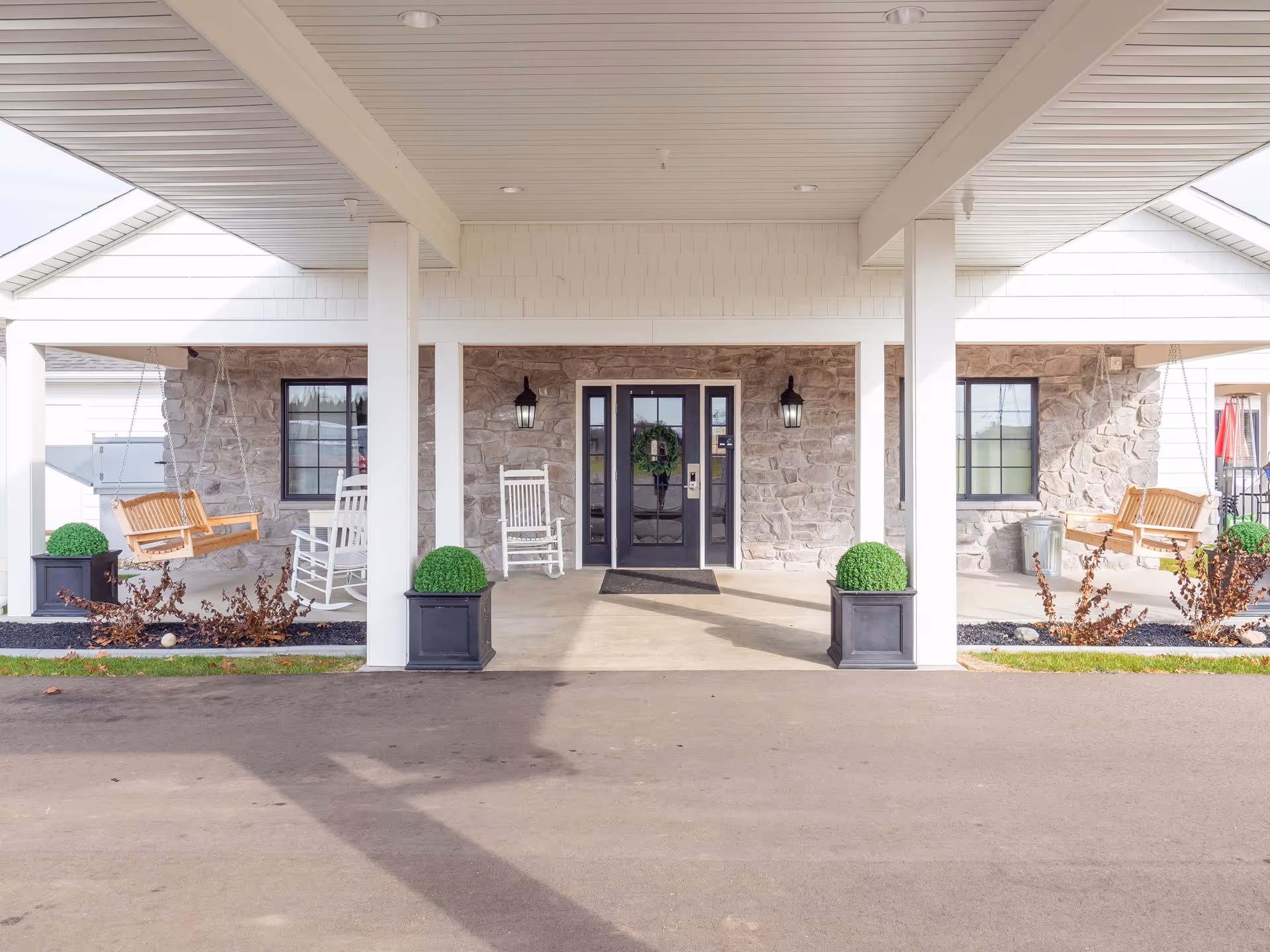 Covered entrance of Birch Meadows Senior Care with a stone wall facade, black double doors with glass panels, two black wall lanterns, white rocking chairs, wooden porch swings on both sides, and decorative green plants in black planters.
