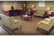 A cozy living room with beige walls and carpet, featuring two armchairs, two sofas, a wooden coffee table with a candle centerpiece, a grandfather clock, a small cabinet, and two table lamps near the windows.