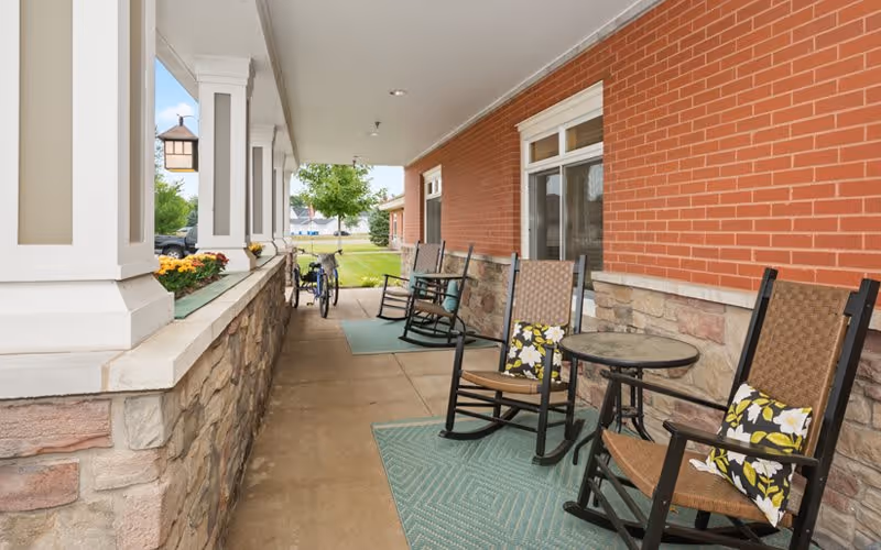 Covered outdoor patio area with several rocking chairs and small round tables on green rugs, adjacent to a brick and stone building wall. A bicycle is parked near the end of the patio, and there is a grassy area with trees visible in the background.