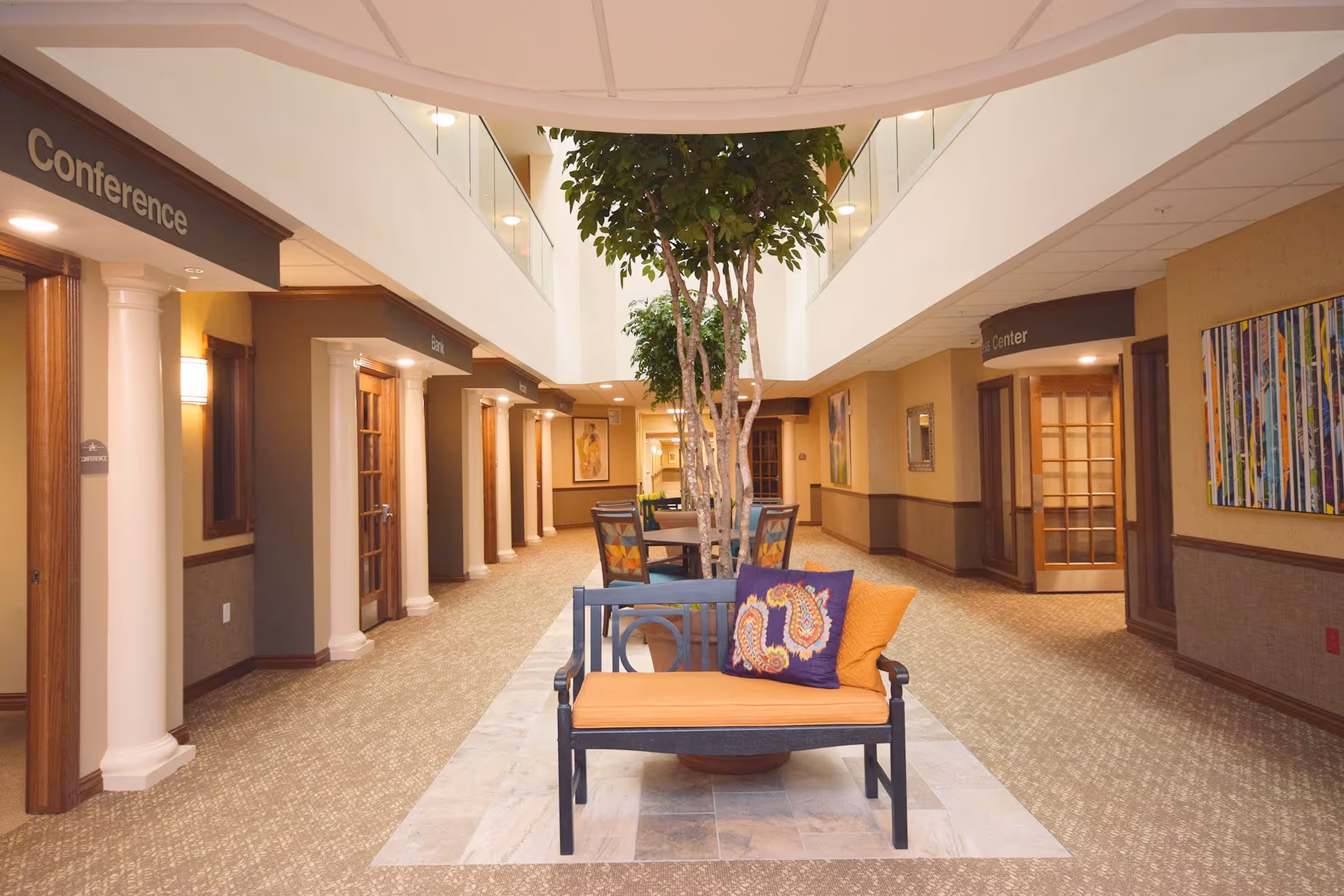Interior hallway of Vista Park Retirement Community featuring a central seating area with a bench adorned with orange and patterned pillows, a large potted tree, and tables with chairs. The hallway is flanked by doors labeled Conference, Bank, and Fitness Center, with warm lighting and decorative artwork on the walls.