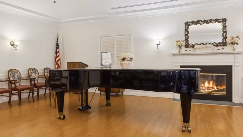 A polished black grand piano in a communal room with a lit fireplace, chairs, floral decor, and an American flag.