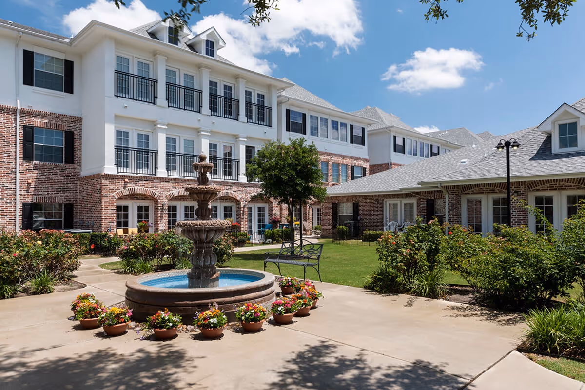 Outdoor courtyard area of a senior living facility with a multi-tiered water fountain surrounded by flower pots, benches, and well-maintained landscaping. The building in the background has a brick and white exterior with multiple windows and balconies under a partly cloudy blue sky.