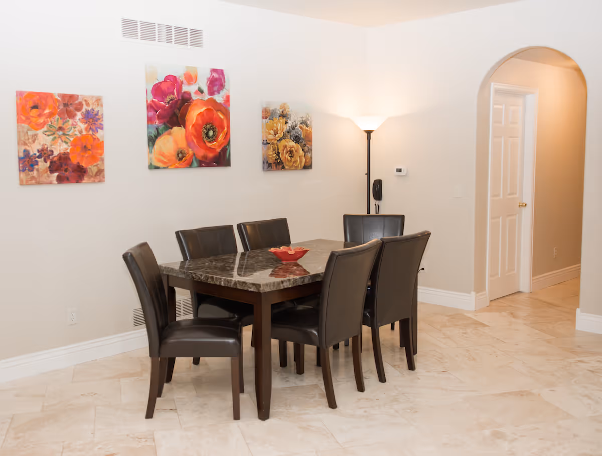 A dining area with a rectangular marble-top table surrounded by six dark leather chairs. Three colorful floral paintings hang on the wall above the table. A floor lamp stands in the corner near an arched doorway leading to another room with a white door. The floor is tiled with light-colored tiles.