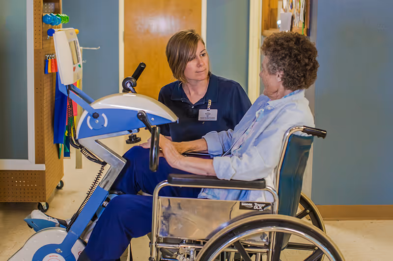 A healthcare worker in a navy blue uniform is attentively talking to an elderly woman seated in a wheelchair. They are in a hallway with light blue walls and a wooden door in the background. The elderly woman is using a rehabilitation exercise machine with pedals.