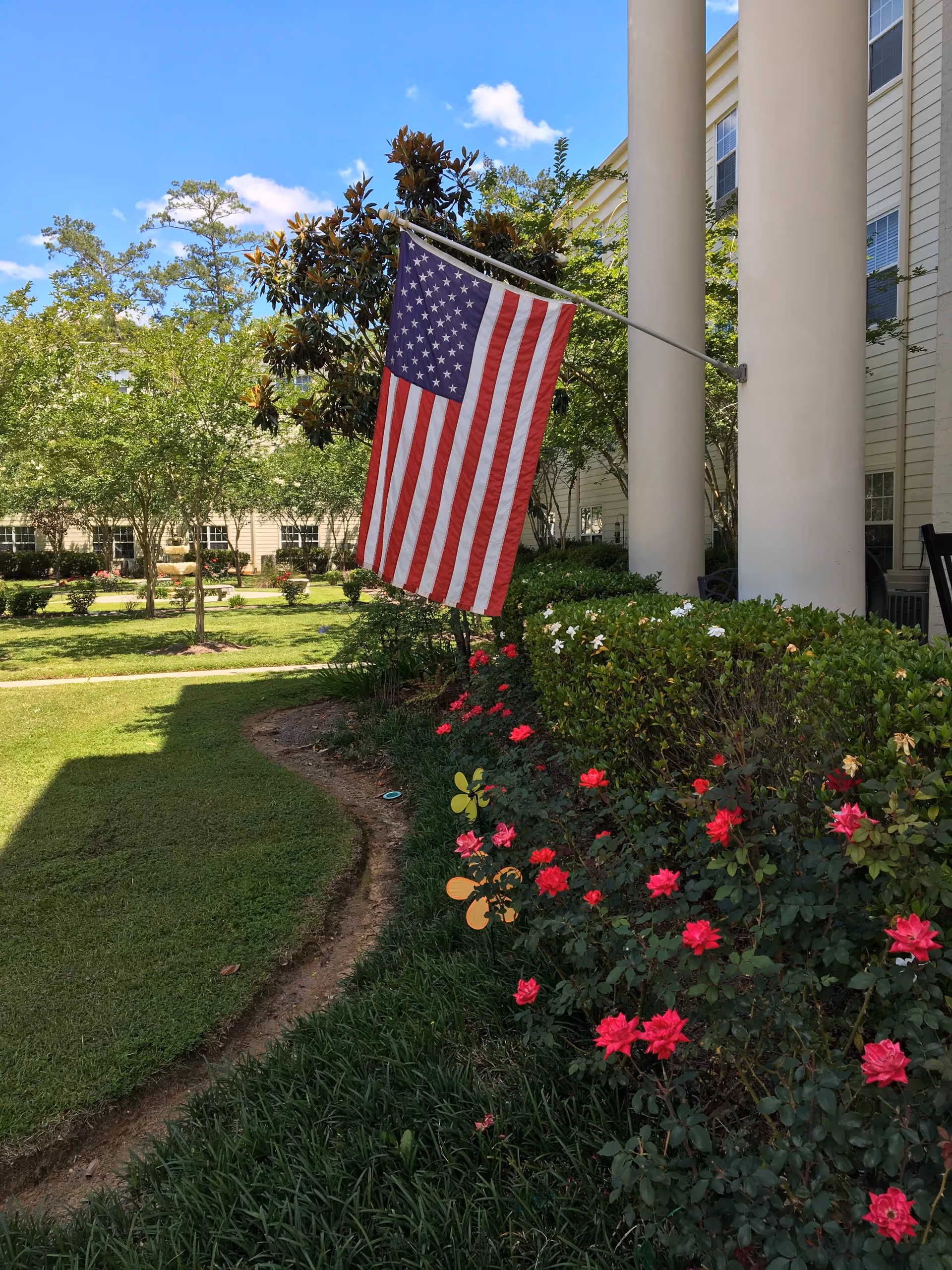 American flag hanging from a pole attached to a building with large white columns, surrounded by green bushes and blooming red and white flowers, with trees and a clear blue sky in the background.