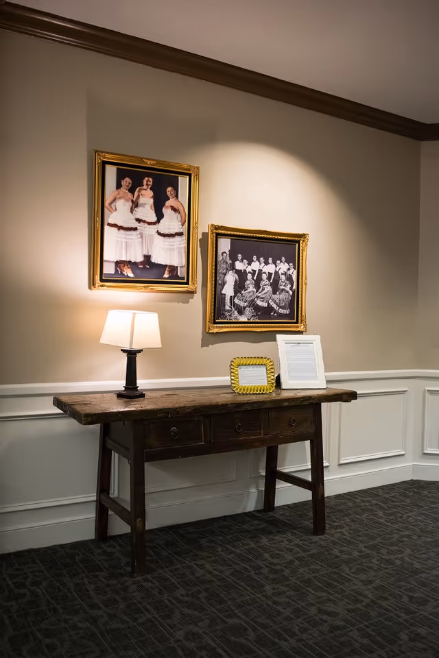 A wooden table with three drawers against a beige wall with white wainscoting. On the table is a small lamp with a white shade, a yellow picture frame, and a white framed document. Above the table are two framed photographs: one of three women in white dresses and another black and white group photo of people in traditional attire.