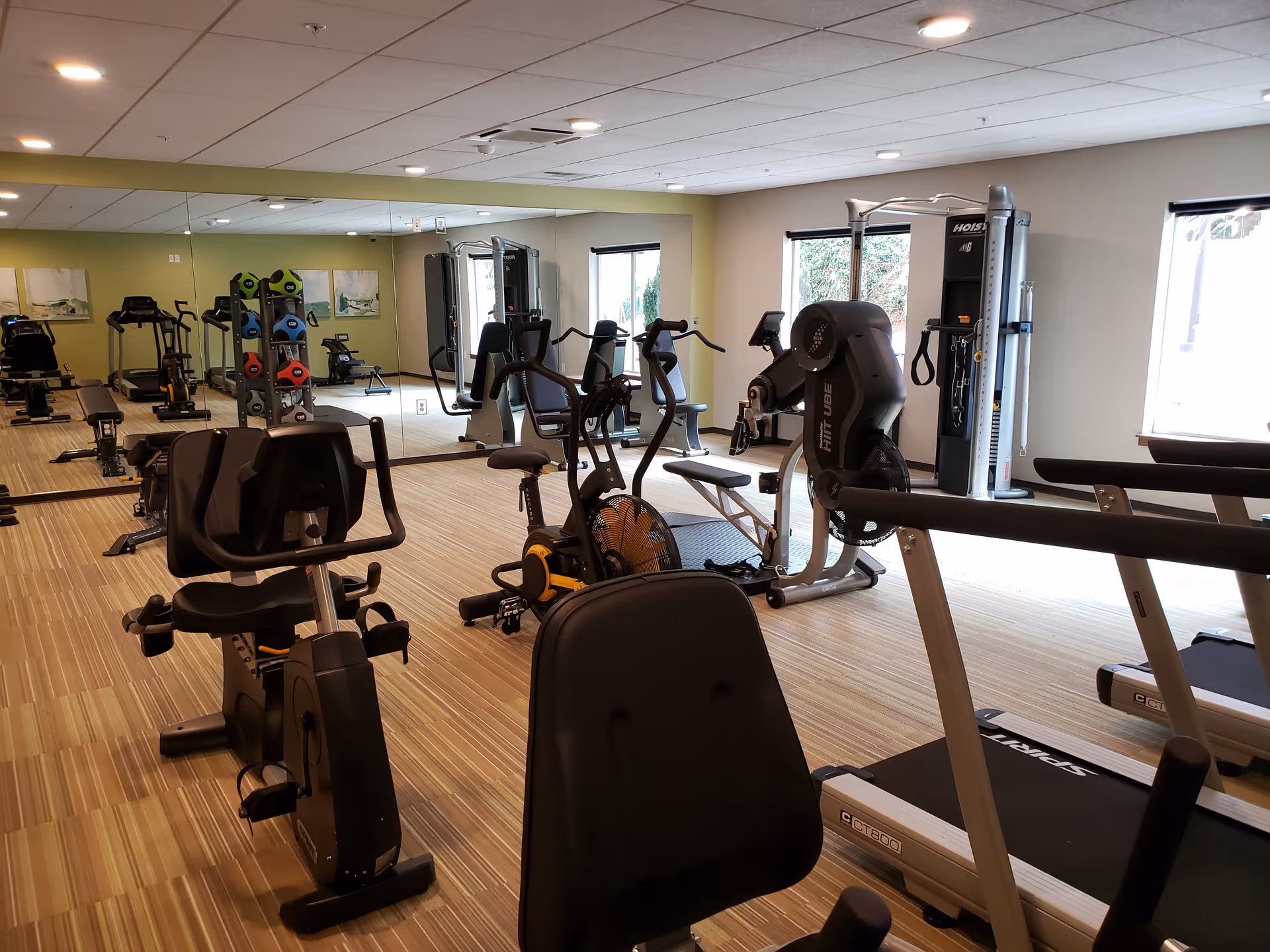 A well-lit fitness room with various exercise equipment including stationary bikes, treadmills, weight machines, and a rack of colorful medicine balls. The room has large windows allowing natural light and a wall covered with mirrors.