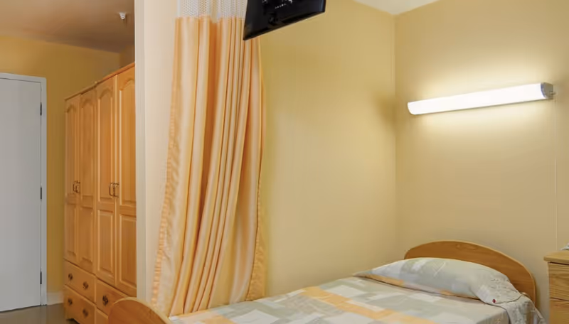 A simple hospital room with a single bed covered with a patterned bedspread, a wooden wardrobe, a wall-mounted light fixture, and a television mounted on the wall. The room has beige walls and a privacy curtain partially drawn.