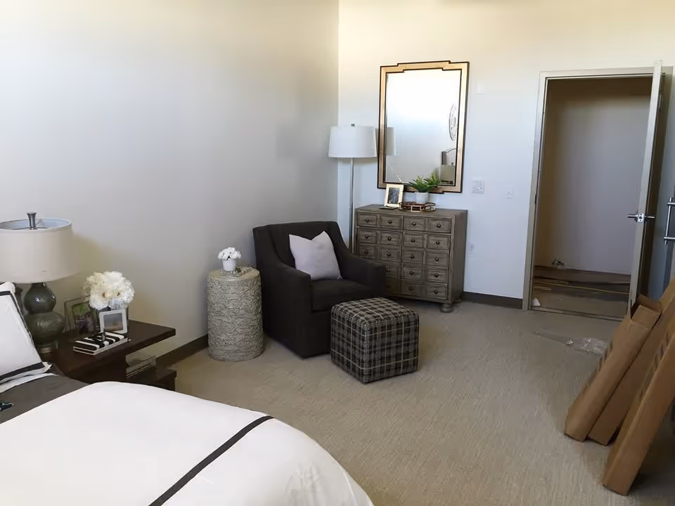 A bedroom with a bed partially visible on the left side, a nightstand with a lamp and decorative items, a dark armchair with a pillow, a plaid ottoman, a round side table with a small flower arrangement, a wooden dresser with a mirror above it, and an open doorway leading to another room. There are some cardboard boxes leaning against the wall on the right side.